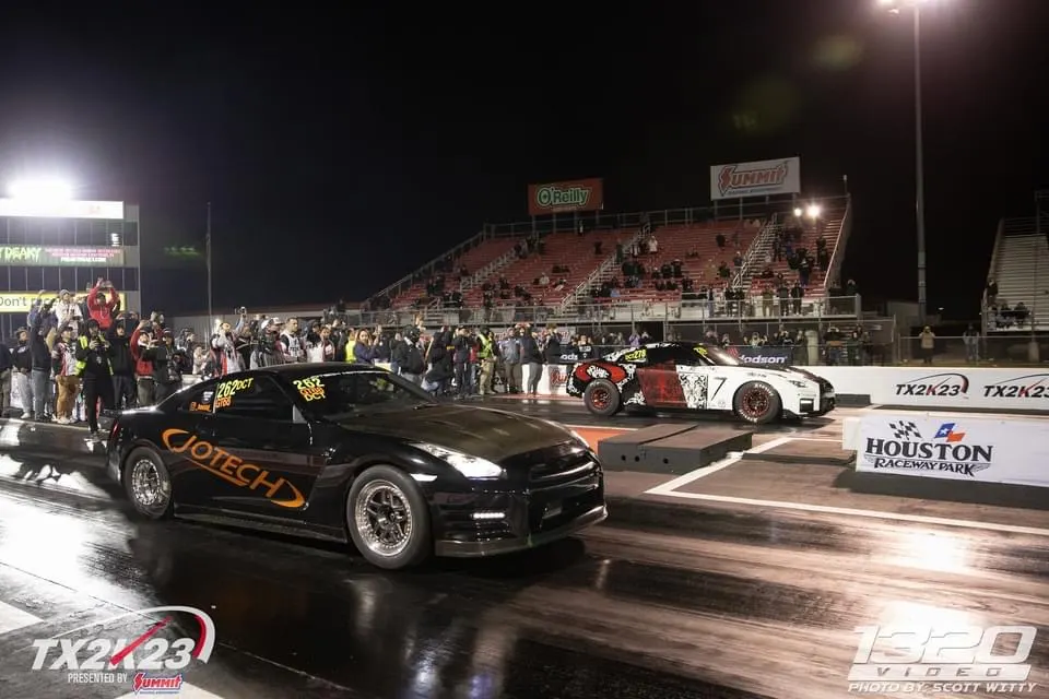 Two high-performance race cars, including a black Nissan GT-R, staged at the starting line of a drag strip at night during TX2K23.