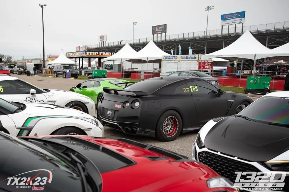 High-performance cars including a Nissan GT-R, Lamborghini Huracan, and Audi R8 parked at the TX2K23 drag racing event paddock.