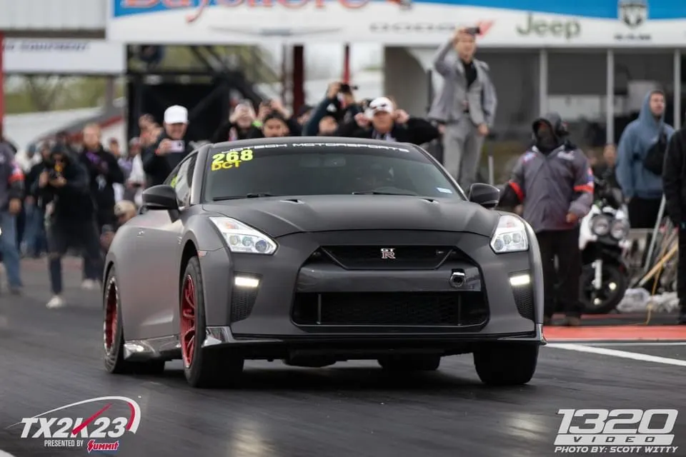 Matte grey Nissan GT-R with red wheels launching on a drag strip at TX2K23, with spectators in the background.