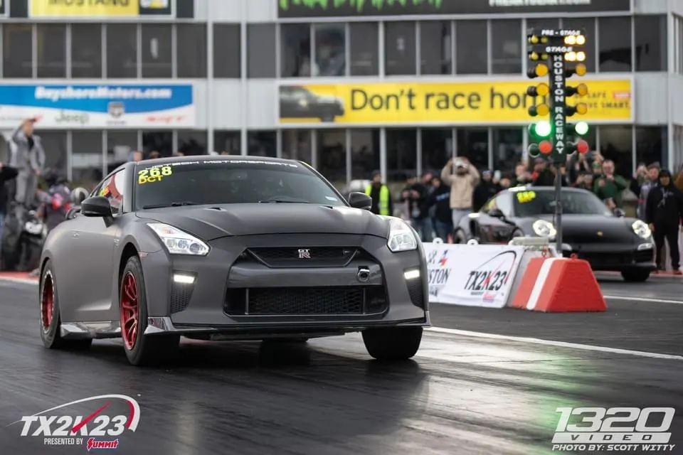A matte grey Nissan GT-R with red wheels launches down a drag strip at TX2K23, with a Porsche in the background.