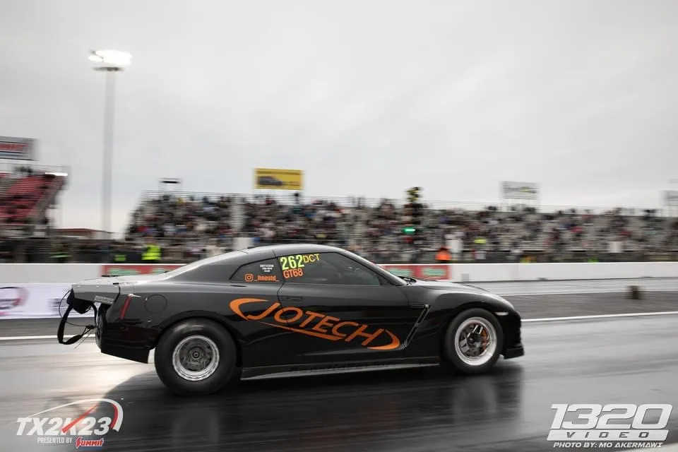 Black Jotech Nissan GT-R drag racing at TX2K23 event on a wet track with spectators in the background.