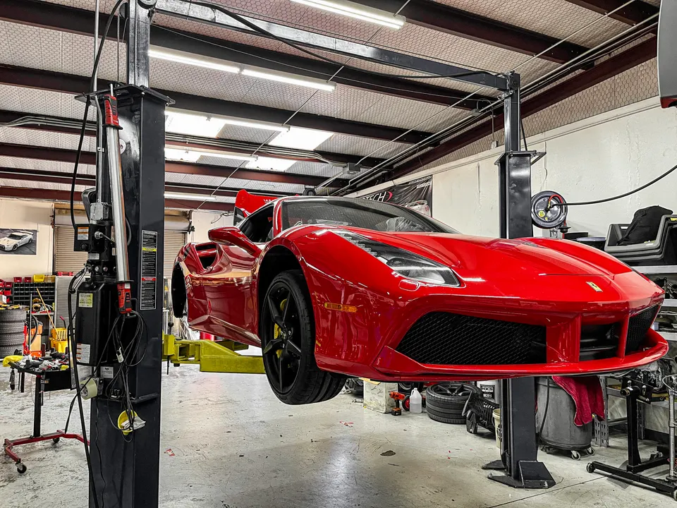 A red Ferrari sports car is elevated on a car lift inside a well-lit automotive performance shop.
