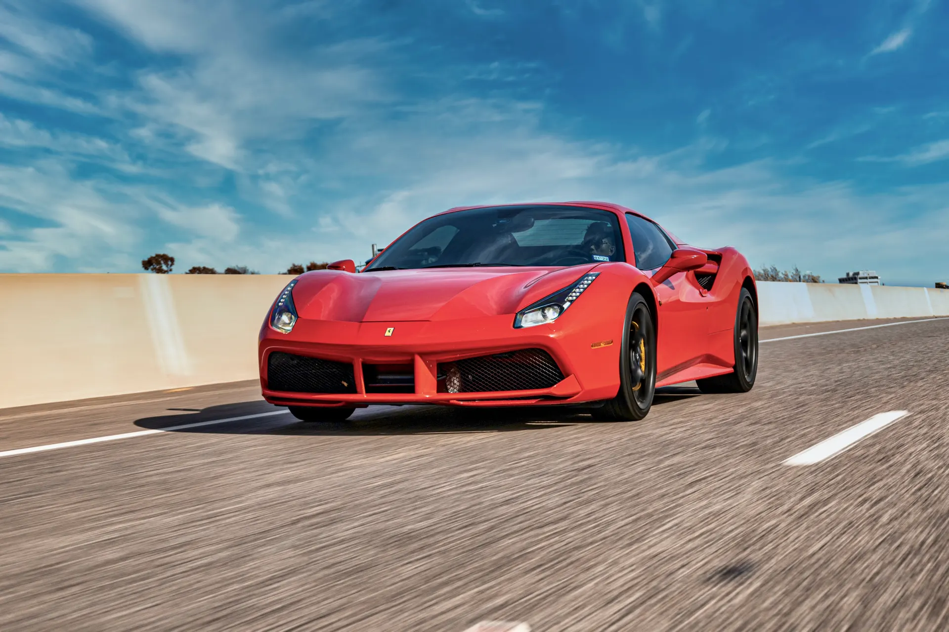 Red Ferrari 488 sports car driving on a highway under a blue sky, showcasing its sleek design and speed.