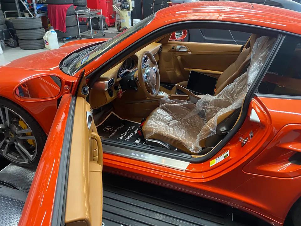 An orange Porsche 911 with its door open, revealing a tan interior with a laptop on the passenger seat in a shop.