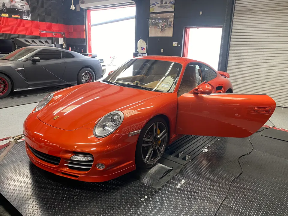 Orange Porsche 997 on a dyno at Jotech Motorsports, with a grey Nissan GT-R in the background.