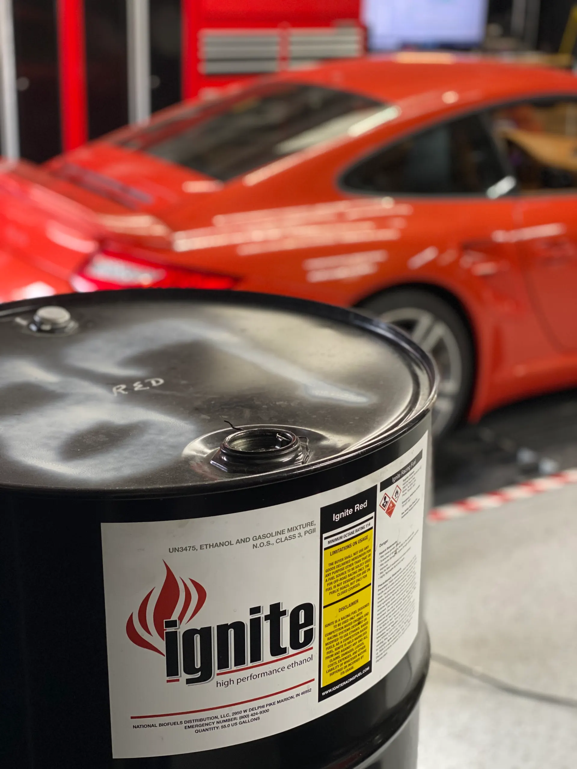 Ignite Red high performance ethanol fuel barrel in a shop with a red Porsche in the background.