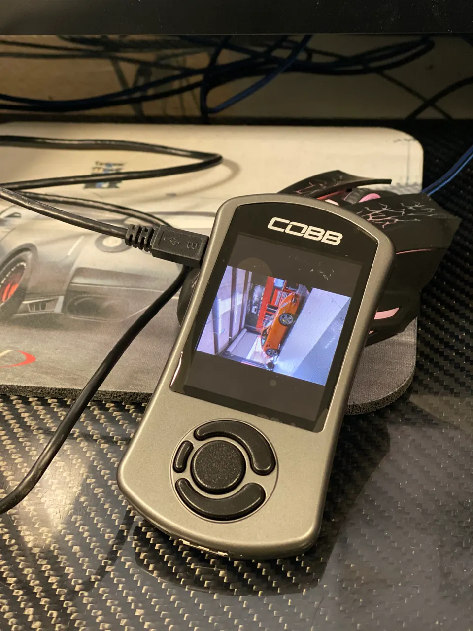 Cobb Accessport tuning device connected via USB on a carbon fiber desk, displaying a car on its screen.
