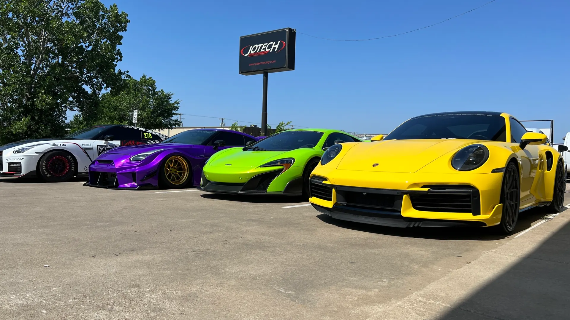 A line of high-performance cars including a white Nissan GT-R, purple Nissan GT-R, lime green McLaren, and yellow Porsche 911 parked at Jotech Motorsports.