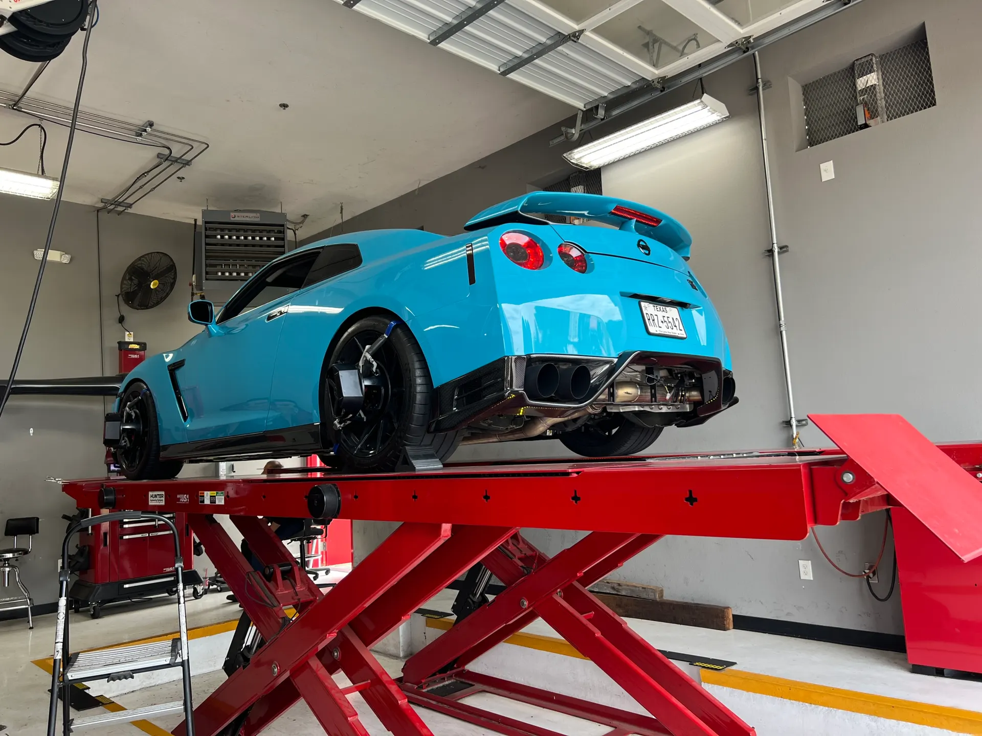 A blue Nissan GT-R on a red car lift in a shop, showing its rear and custom exhaust system during an alignment.