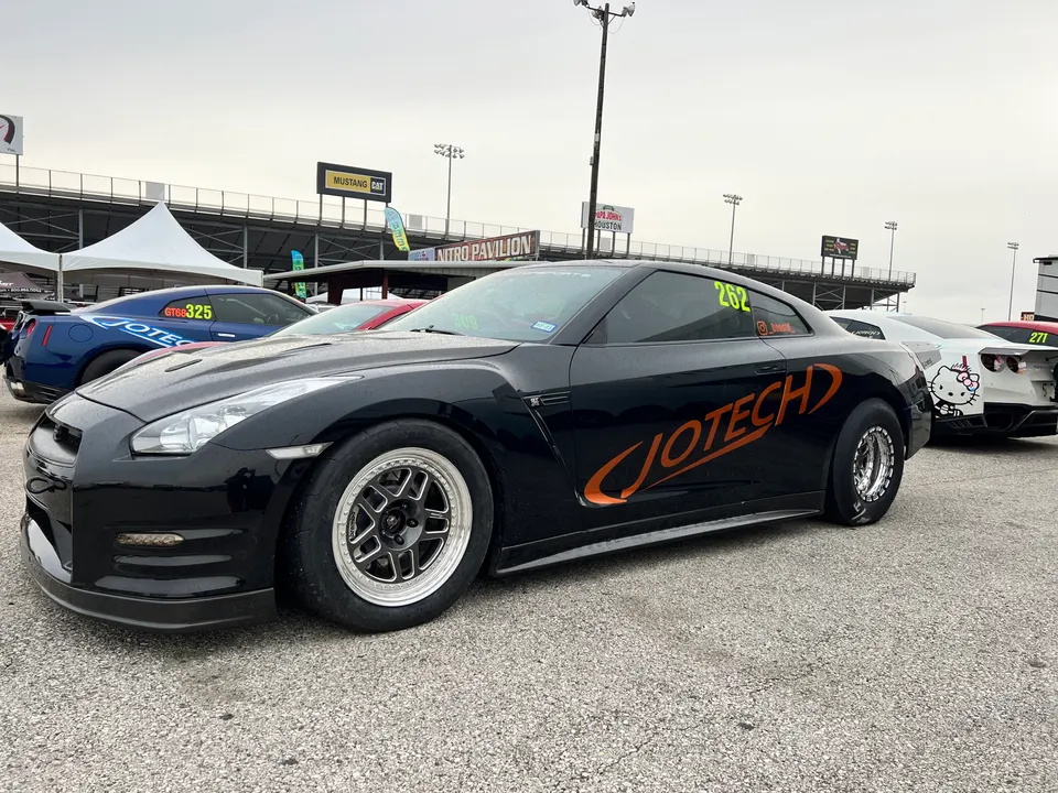 A black Nissan GT-R with Jotech livery and drag wheels parked at a drag strip with other cars and tents.