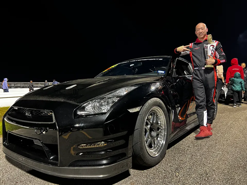 A black Nissan GT-R on a drag strip at night with a driver in a race suit holding a trophy.