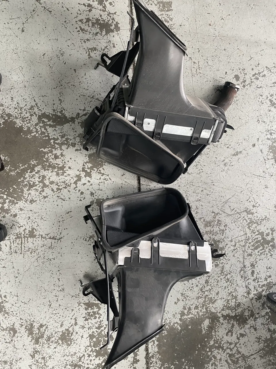 Two black plastic air intake boxes and ducts on a concrete shop floor, likely removed from a performance car.