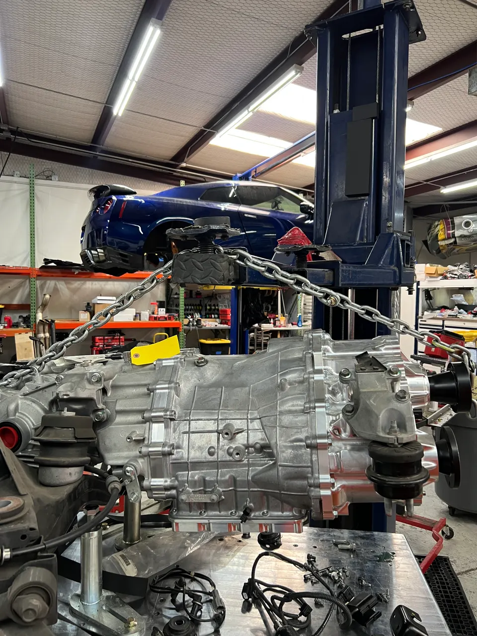 Nissan GT-R on a car lift in a shop with a transmission assembly suspended in the foreground, ready for installation.
