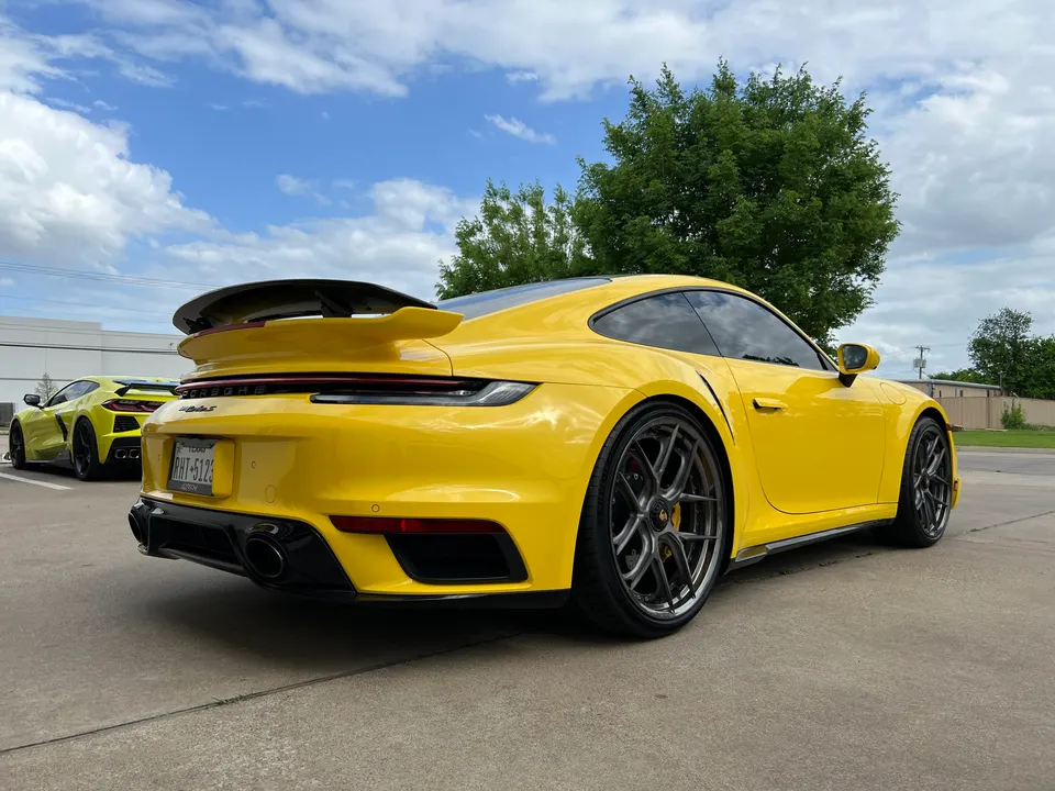 Yellow Porsche 911 with a large rear wing and custom wheels parked outdoors, with a yellow Lamborghini in the background.