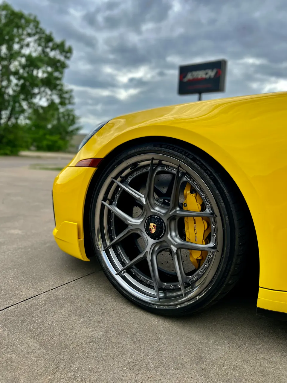Close-up of a yellow Porsche's custom multi-spoke wheel and yellow brake caliper, with a Jotech sign in the background.