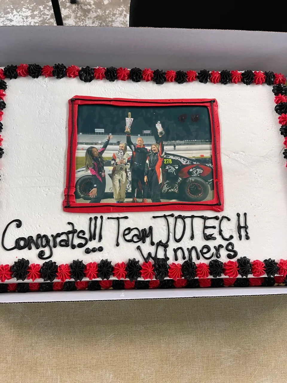 A celebratory cake for Team JOTECH Winners, featuring an edible image of the team with trophies and a race car on a track.
