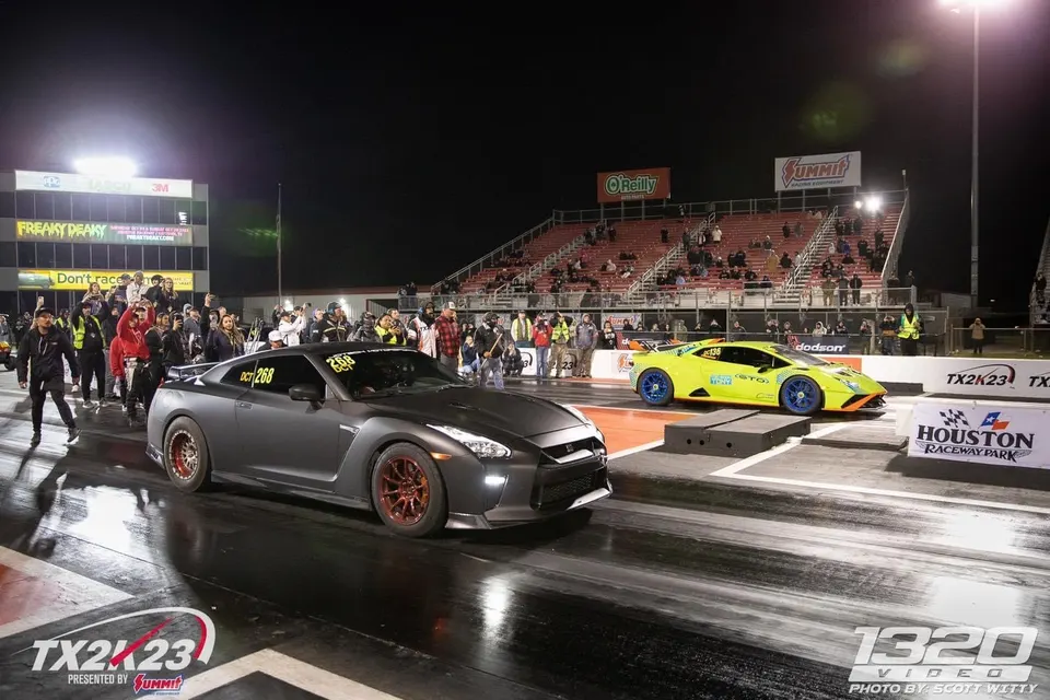 A matte grey Nissan GT-R and a neon yellow Lamborghini Huracan at the starting line of a drag strip at night during TX2K23.