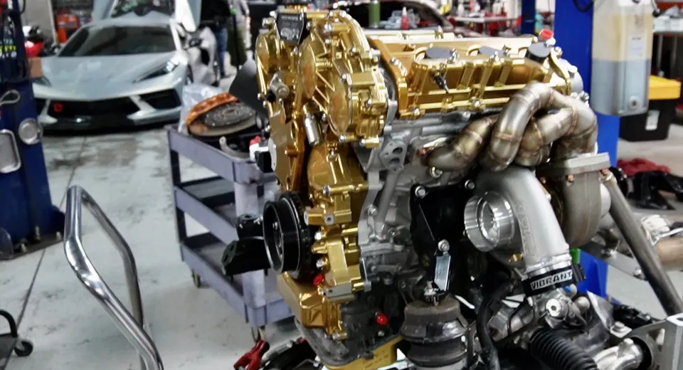 Custom gold engine with a large turbocharger and custom exhaust manifold being built in a performance shop, with a C8 Corvette in the background.