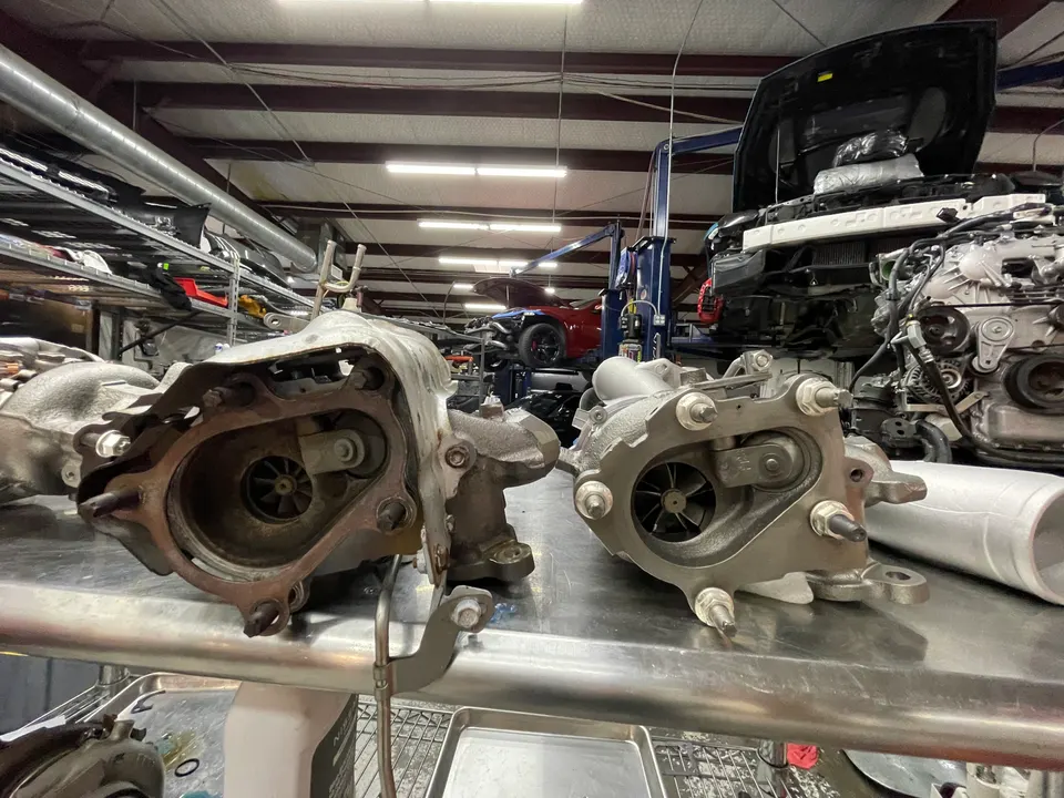 Two turbochargers on a metal workbench in a performance tuning shop, with a car on a lift in the background.