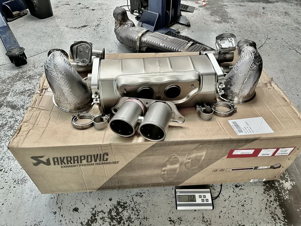 Akrapovic exhaust system with heat shields and clamps on a cardboard box in a performance shop, showing weight on a scale.