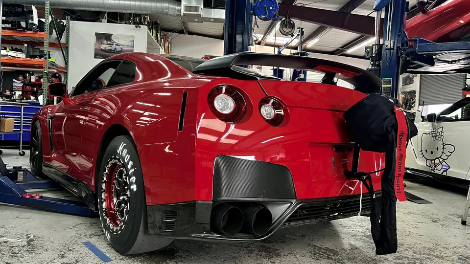 Red Nissan GT-R drag car with large rear wing, carbon diffuser, and parachute in a performance tuning shop.