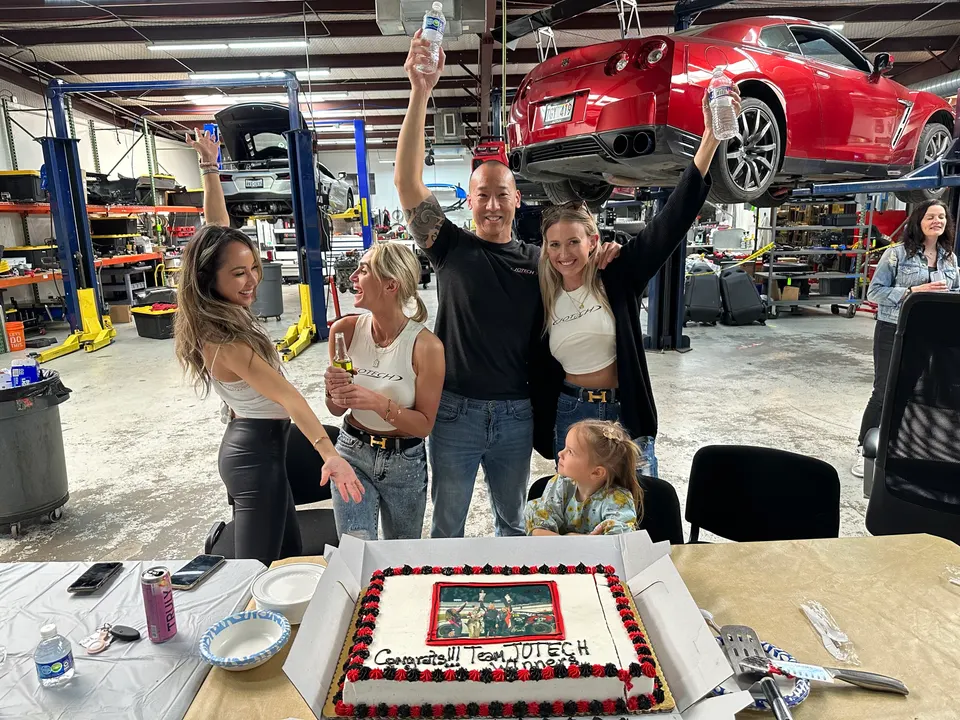 Jotech Motorsports team celebrating in the shop with a cake, a red Nissan GT-R and a silver Corvette on lifts in the background.