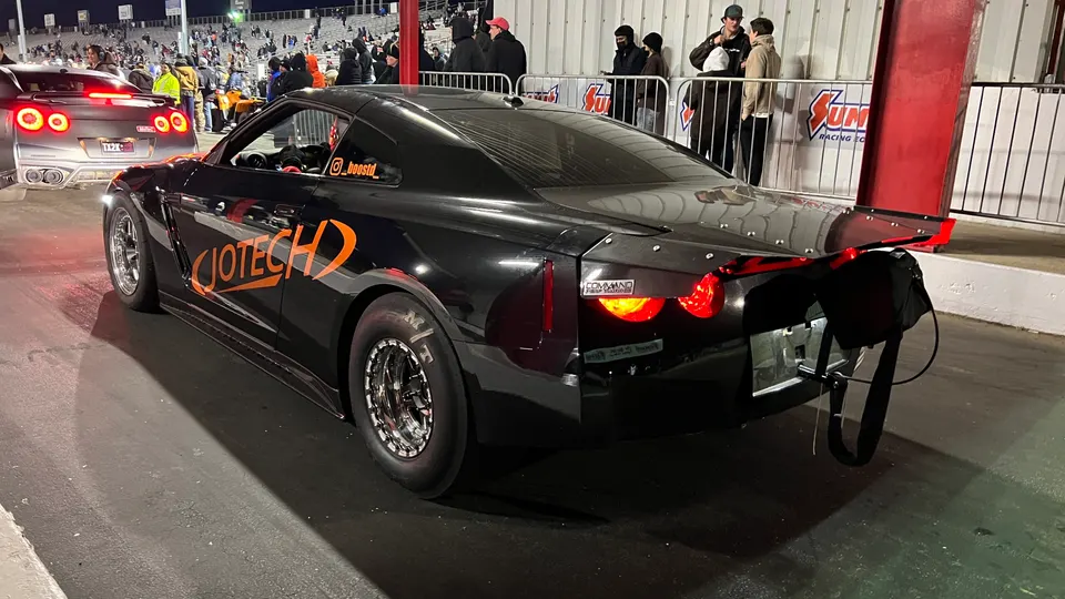 A black Jotech Nissan GT-R drag car with a parachute and large rear wing at a drag strip, with another GT-R in the background.