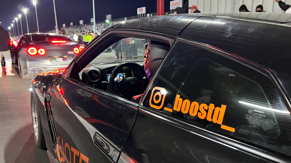 A black Nissan GT-R with an orange '_boostd_' sticker on the window waits in the staging lane at a drag strip at night, with another GT-R in the background.