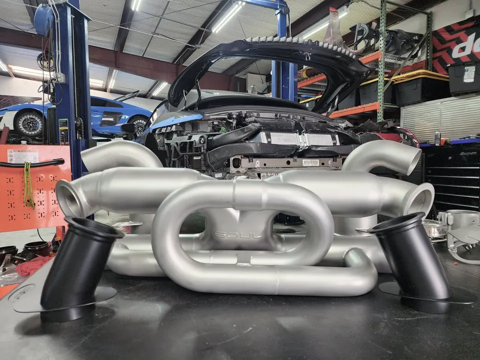 A Soul Performance exhaust system on a workbench in a tuning shop with an Audi R8 on a lift in the background.
