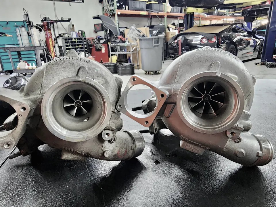 Two large turbochargers on a workbench in a performance tuning shop, with a Nissan GT-R in the background.