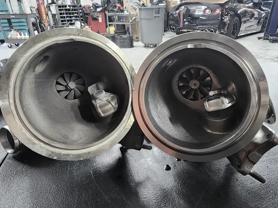 Two large turbochargers with internal wastegates displayed on a workbench in a performance tuning shop, with a Nissan GT-R in the background.
