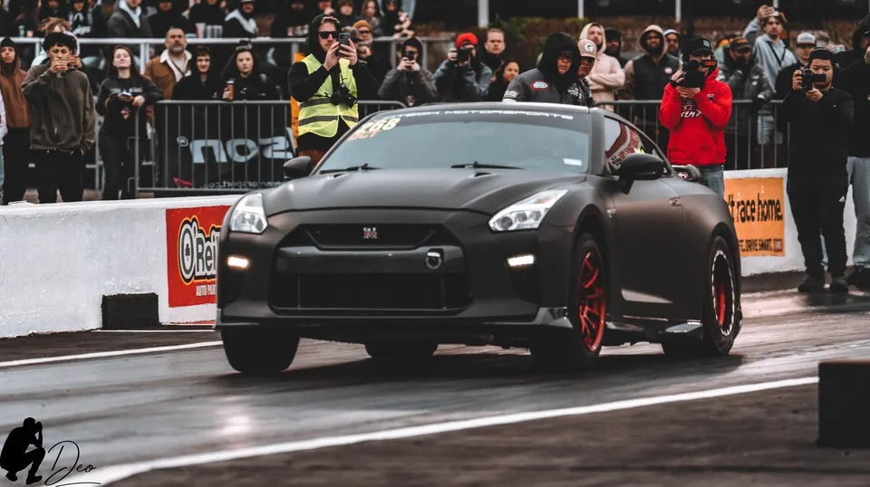 A matte black Nissan GT-R with red wheels on a drag strip, with spectators watching in the background.