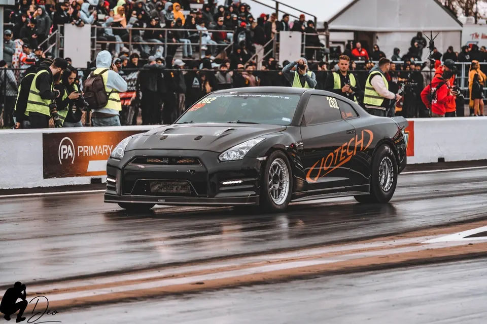 A black Jotech Motorsports Nissan GT-R on a wet drag strip with spectators in the background, ready for a race.
