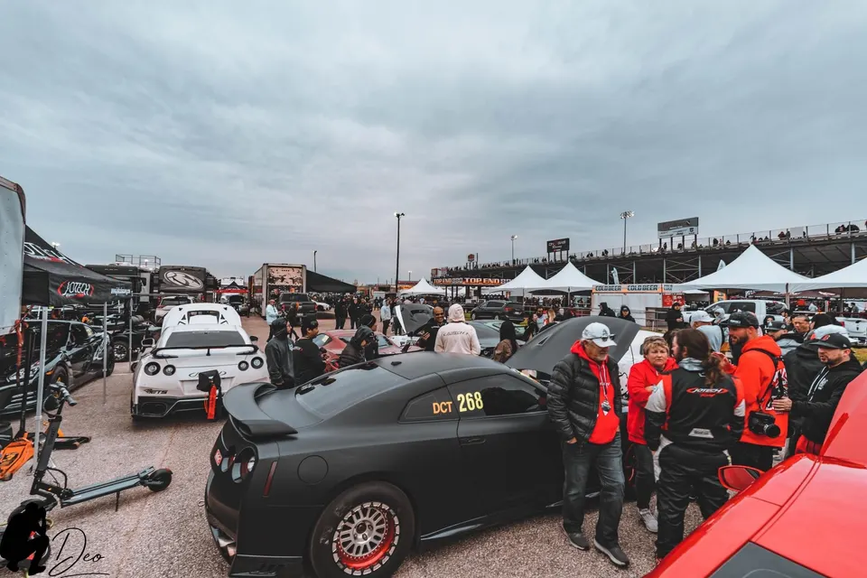 Crowd gathered around modified Nissan GT-Rs and other performance cars in the paddock area of a drag racing event.