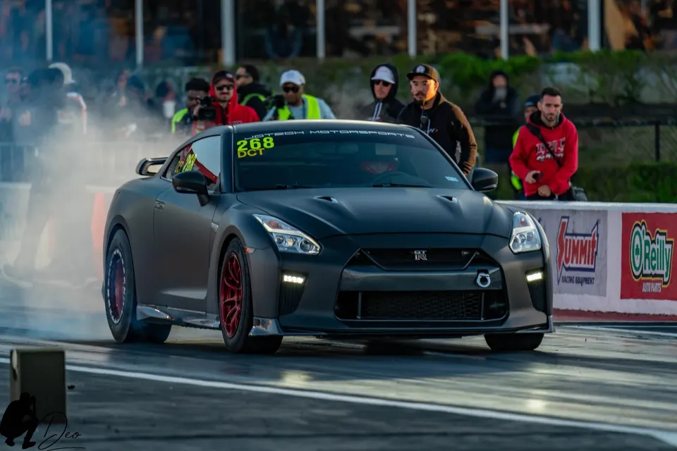 A matte black Nissan GT-R R35 performs a burnout on a drag strip, with red aftermarket wheels and spectators in the background.
