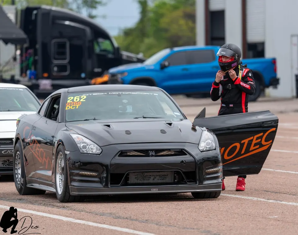 A black Nissan GT-R race car with a driver in a racing suit and helmet standing next to it at a drag strip staging lane.