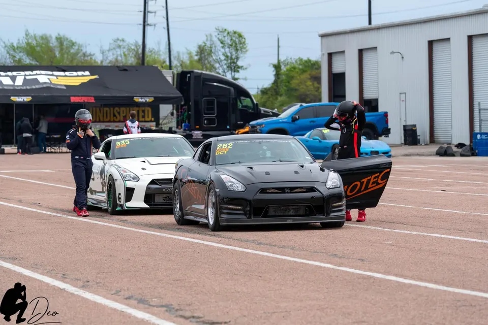 Two Nissan GT-R race cars, one white and one black, with drivers in racing suits at a drag strip staging lane.