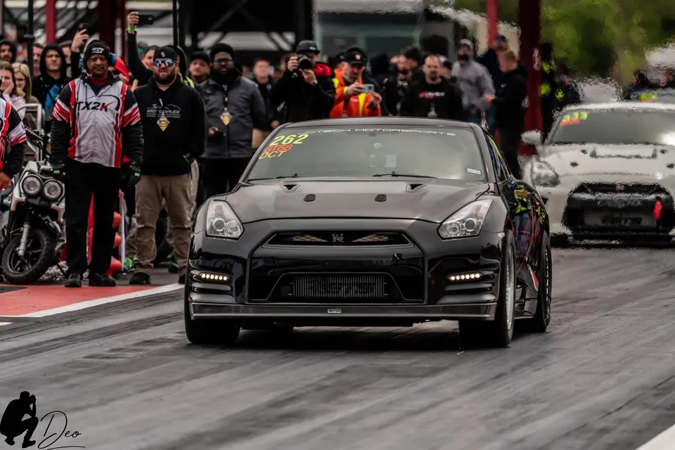 A black Nissan GT-R race car on a drag strip, with spectators and another GT-R in the background.