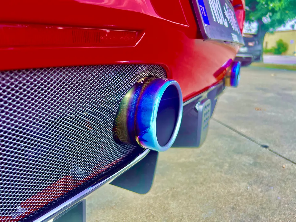 A close-up of a red car's rear bumper featuring a vibrant blue and purple titanium exhaust tip and a mesh grille.