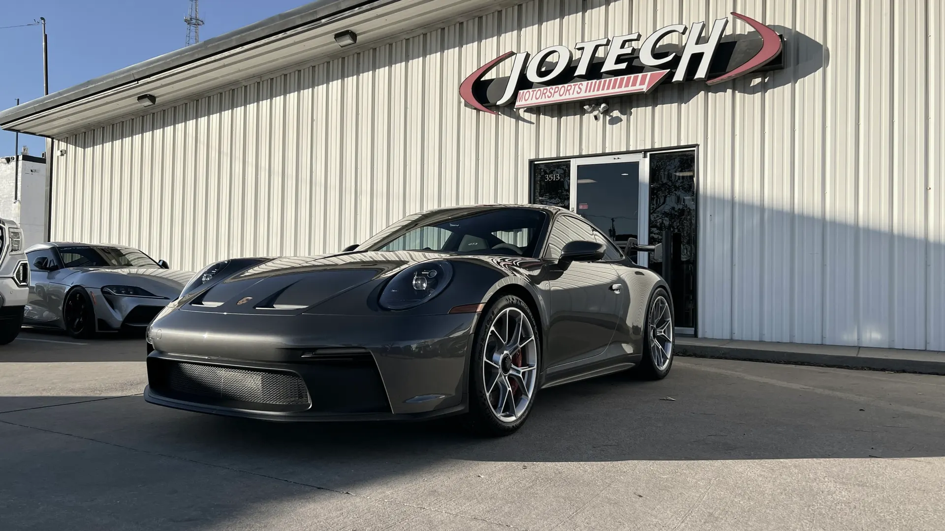 Grey Porsche 911 GT3 parked outside Jotech Motorsports performance shop with a silver Toyota Supra in the background.