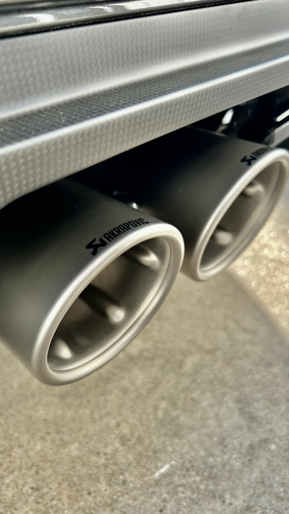 Close-up of matte grey Akrapovic exhaust tips on a car, with a carbon fiber textured bumper visible above.