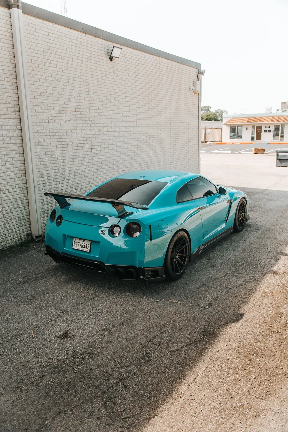 A turquoise Nissan GT-R with a carbon fiber wing and custom wheels parked next to a white brick building.