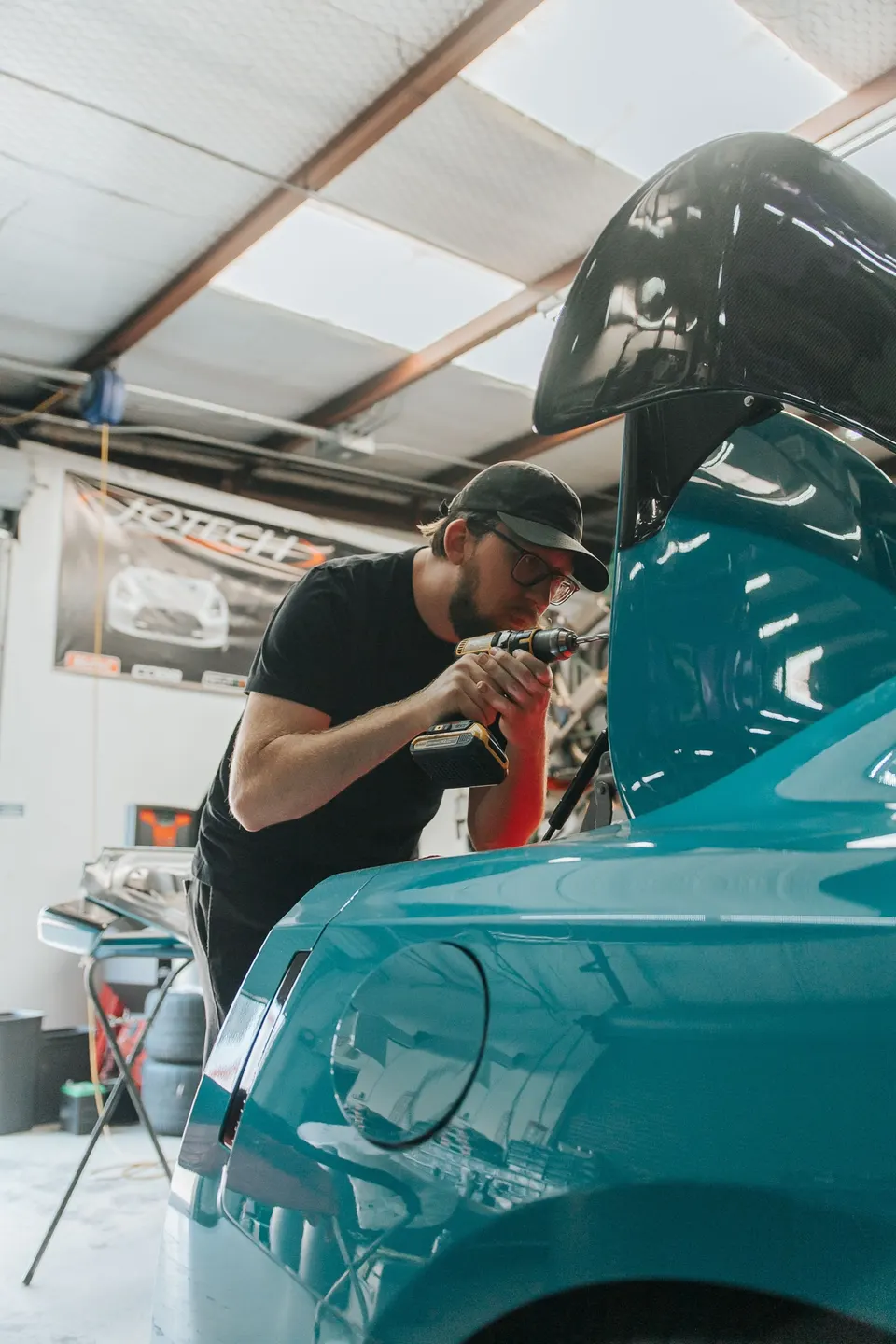 Mechanic drilling into the rear of a teal sports car to install a large carbon fiber wing in a performance tuning shop.