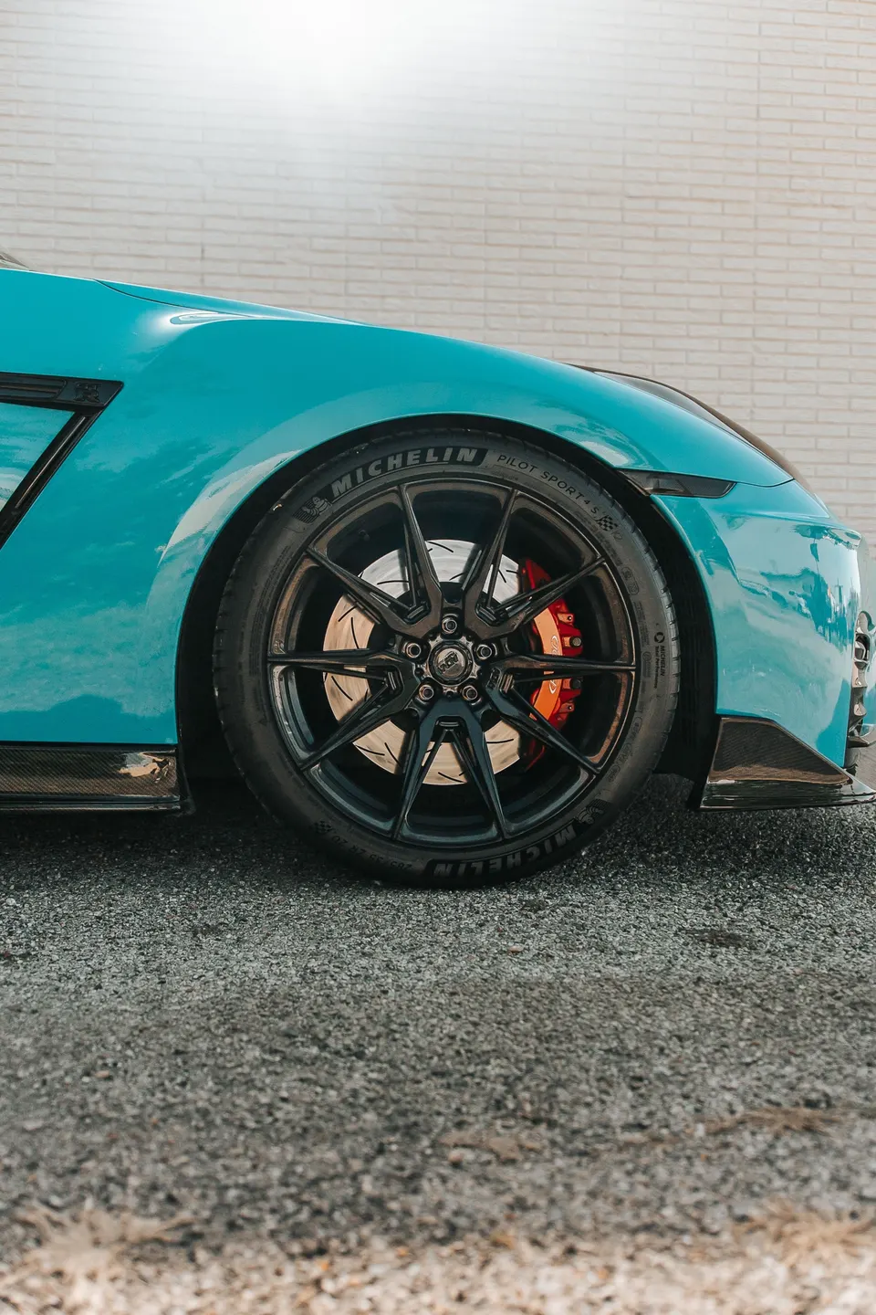 A teal Nissan GT-R's front wheel with black multi-spoke rims, Michelin tires, and red brake calipers against a brick wall.