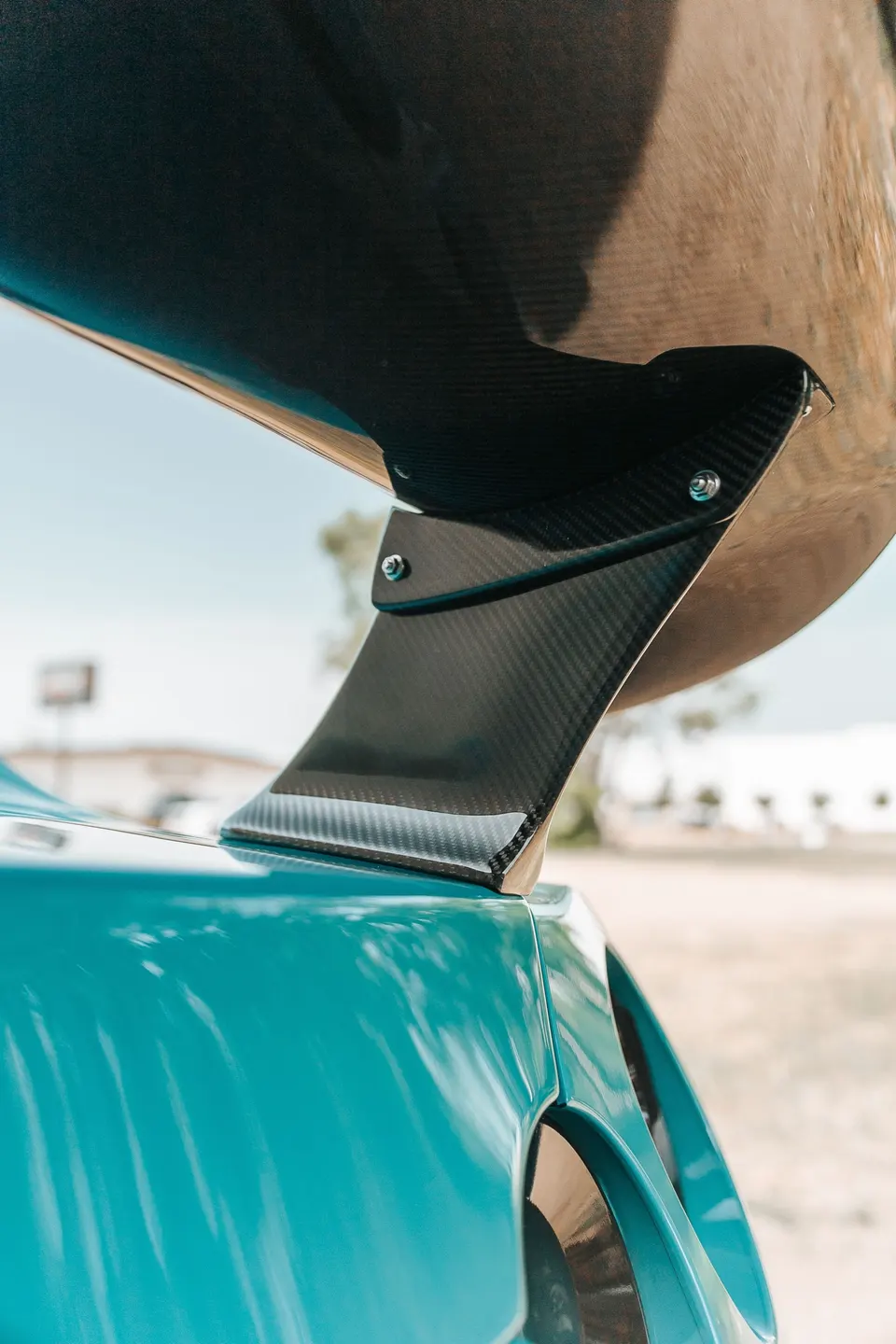 Close-up of a carbon fiber rear wing on a teal sports car, showcasing the aerodynamic detail.