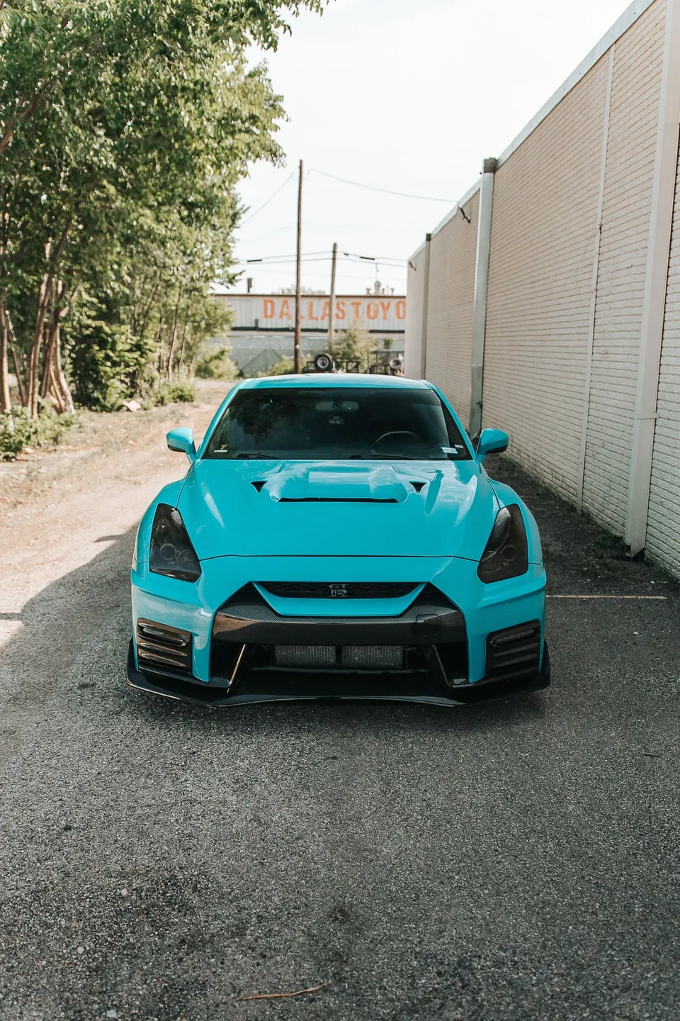 Turquoise Nissan GT-R with a carbon fiber front splitter and custom headlights parked in an alley.