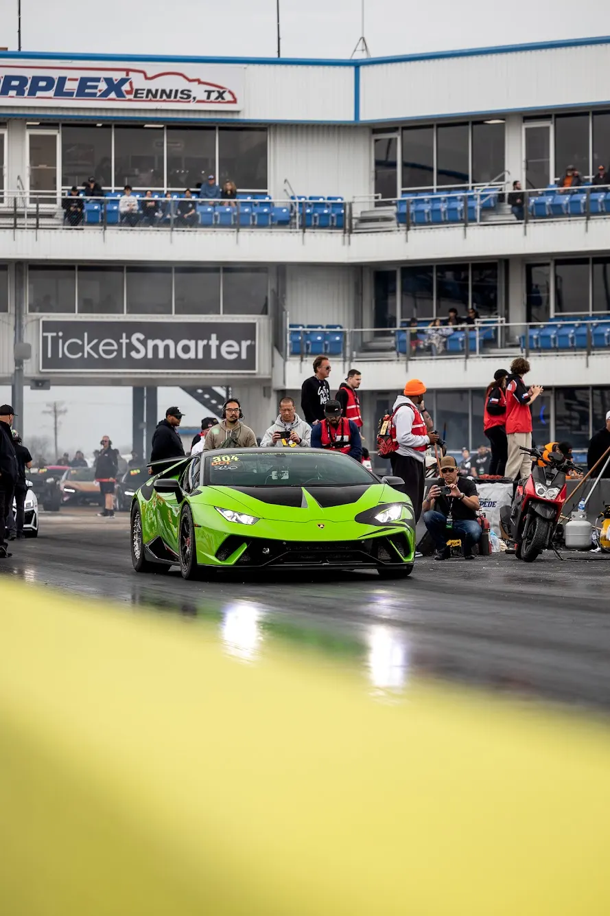 A bright green Lamborghini Huracan in the staging lanes of a drag strip at Texas Motorplex, with spectators and buildings in the background.