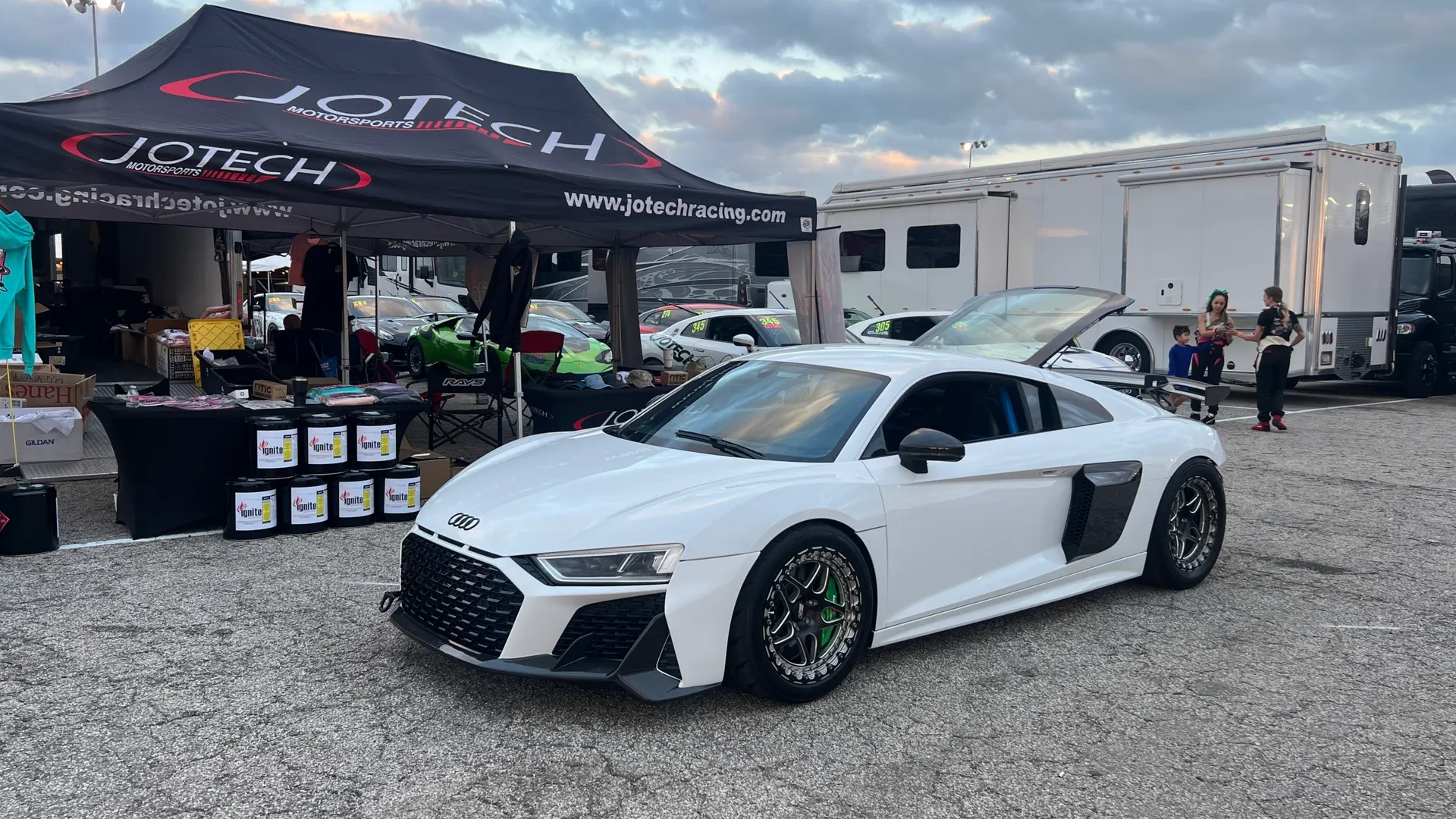 White Audi R8 supercar parked in a Jotech Motorsports paddock area at a track event with trailers and people in the background.