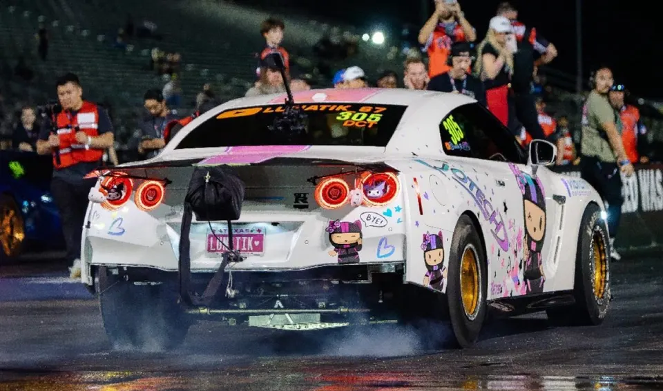 A white Nissan GT-R drag car performs a burnout on a drag strip at night, with spectators in the background.