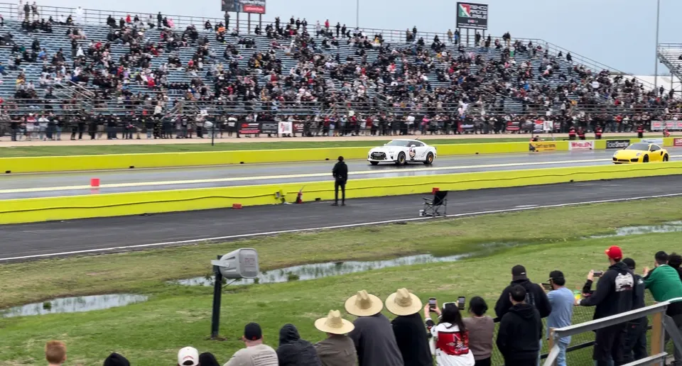 Two high-performance cars, a white Nissan GT-R and a yellow Porsche, racing down a drag strip with spectators in the stands.